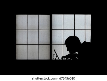 Window silhouette of a creepy computer hacker hunched over a laptop late at night