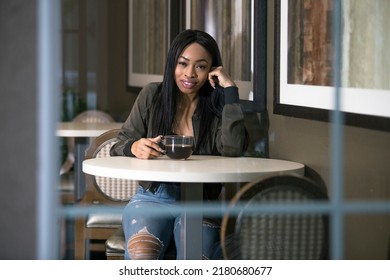 Window view of a black female having coffee in a coffeeshop or sidewalk cafe.  The entrepreneur businesswoman on a break or student is waiting patiently and looks independent.