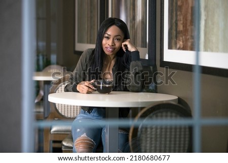Window view of a black female having coffee in a coffeeshop or sidewalk cafe.  The entrepreneur businesswoman on a break or student is waiting patiently and looks independent.
