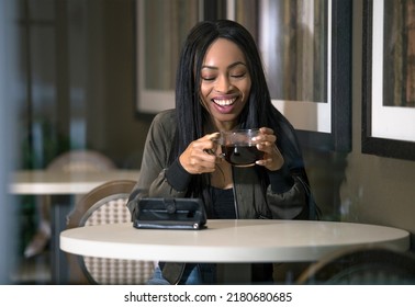 Window View Of A Black Female Watching Streaming Videos On A Cell Phone In A Coffeeshop Via 5g Wifi Internet.  She Is Sitting And Holding A Cup Of Coffee.