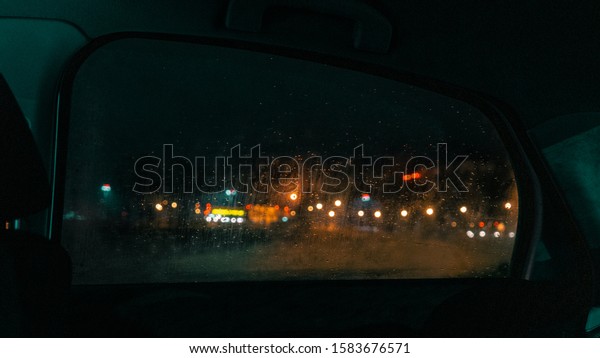 Window view of a car on a rainy night. The glass is\
foggy and drops are sliding on it, in the distance the lights of\
the night city.