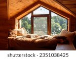 A window with a wide cozy window sill on the second floor of a wooden house. The window sill is covered with a fur blanket and pillows. The window overlooks the forest.