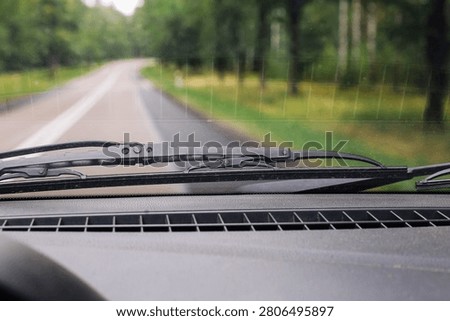 Windshield wipers work during rainy drive on country road. Wet car window background shows blurred forest and asphalt ahead. Vehicle travel scene represents bad weather road safety and visibility.