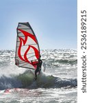 Windsurfer and his bold graphic red and black windsail as he surmounts a wave at Jalama Beach, California