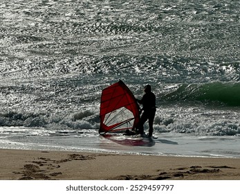 A windsurfer prepares to enter the ocean, holding a red sail while standing at the water’s edge. The shimmering waves and sandy beach create a dynamic scene, highlighting the adventurous spirit of wat - Powered by Shutterstock