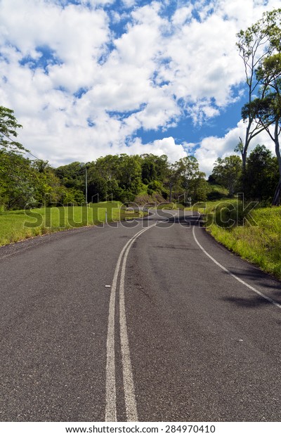 Windy asphalt road leading uphill. Green grass and trees\
either side of road. Blue sky with white clouds. Vertically\
orientated image. Image taken from centre of road. Image taken in\
rural Australia. 