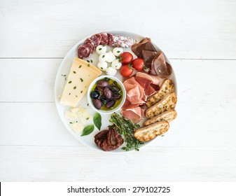 Wine Appetizer Set. Cherry-tomatoes, Parmesan Cheese, Meat Variety, Bread Slices, Dried Tomatoes, Olives And Basil On Round Ceramic Plate Over White Wood Backdrop, Top View