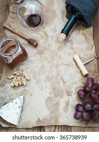 Wine and appetizer set with copy space in center. Glass of red wine, bottle, corkscrewer, blue cheese, grapes, honey and walnuts on oily craft paper over rustic wooden table, top view