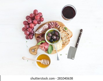 Wine appetizer set. Glass of red wine, grapes, honey, parmesan cheese, meat variety, bread slices, pecan nuts, honey, olives and basil on rustic wooden board over white wood backdrop, top view - Powered by Shutterstock