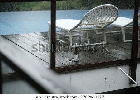 Wine Bottle and Empty Glasses on Wooden Terrace in the Rain with Sunlounger Background