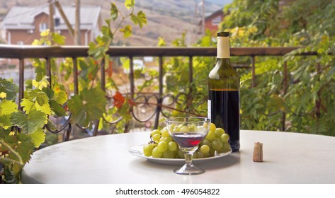 Wine Bottle,wine Glass And Grapes On A Round Table. Balcony,wreathed With Grape Leaves.Crimea. 