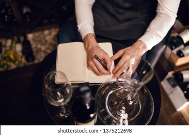 Wine Degustator Man Taking Notes In Winery With Wine Bottles Stacked In Rows On The Shelves. Wine Tester Writing In Notepad Information Of Wine Origin And Taste, Top View.