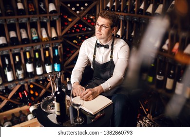 Wine Degustator Man Taking Notes In Winery With Wine Bottles Stacked In Rows On The Shelves. Wine Tester Writing In Notepad Information Of Wine Origin And Taste.