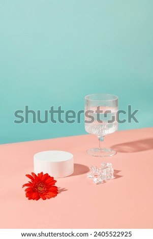 A wine glass containing water, a red gerbera flower, ice and a round white podium stand out against a blue-pink background. Display products on an empty podium.