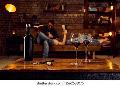 Wine Glasses Of Red Wine On Table At Home Couple Reading Book In Background. Autumn Or Winter Mood With Dark And Warm Colors.