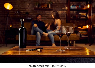 Wine Glasses Of Red Wine On Table At Home Happy Love Couple Talking In Background. Autumn Or Winter Mood With Dark And Warm Colors.