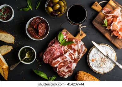 Wine snack set. Glass of red, meat selection, mediterranean olives, sun-dried tomatoes, baguette slices, camembert cheese and spices on black background, top view - Powered by Shutterstock