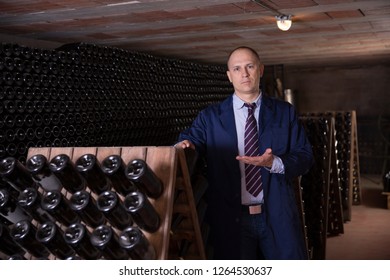 Сonfident winemaker inviting to wine cellar, demonstrating bottles with aging wine on racks