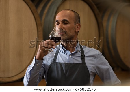 Winemaker smelling a glass of red wine in a cellar surrounded by wooden barrels. Professional mature man smelling red wine in glass with closed eyes in a wine cellar. Sommelier inspecting wine. Сток-фото © 