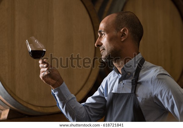 Winemaker in wine\
cellar holding glass of red wine and checking it. Sommelier testing\
wines in winery basement. Wine producer inspecting quality of red\
wine in front of\
barrels.\
