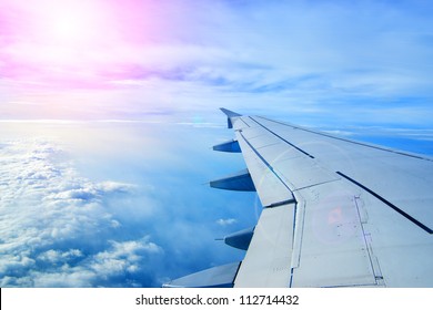 Wing Of An Airplane Flying Above The Clouds. People Look At The Sky From The Window Of The Plane, Using Air Transport To Travel. Back Light Sun Beam