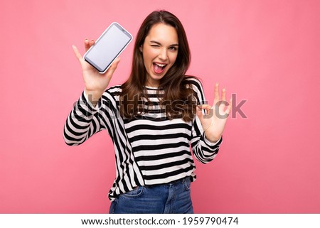 Winking beautiful happy young woman wearing striped sweater isolated over background with copy space showing ok gesture looking at camera showing mobile phone screen display