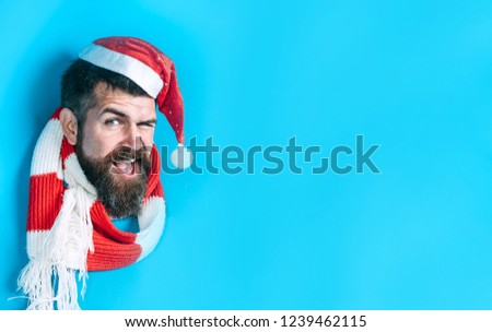 Winking guy in Santa hat breaks through paper wall. Happy man in Santa hat looking through hole in paper. Christmas, New Year, holidays, winter concept. Stylish guy in santa hat&scarf. Christmas sales