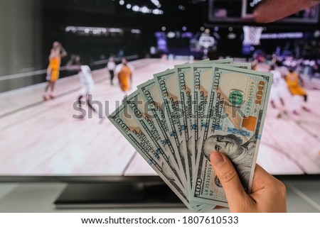 The winner at basketball betting holds a dollar money prize in hand near the TV