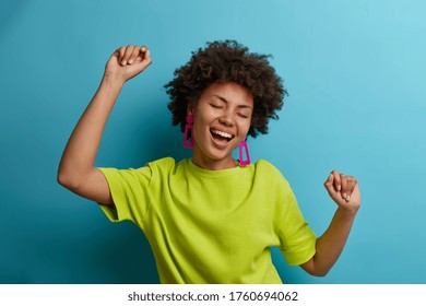 I am winner! Ecstatic overjoyed African American woman dances carefree, celebrates victory and success, dressed in green casual t shirt, feels lively and energetic, isolated on blue background - Shutterstock ID 1760694062