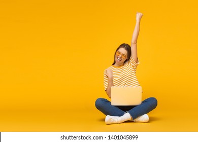 Winner! Excited smiling girl sitting on floor with laptop, raising one hand in the air is she wins, isolated on yellow background - Shutterstock ID 1401045899