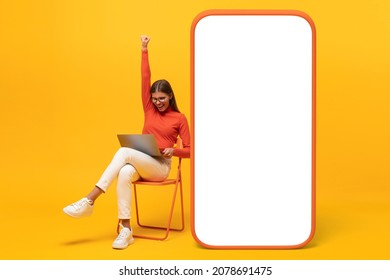 Winner. Excited Young Woman Sitting On Chair With Her Laptop And Huge Phone Mock Up With Blank Screen, Shouting Yes, Isolated On Yellow 