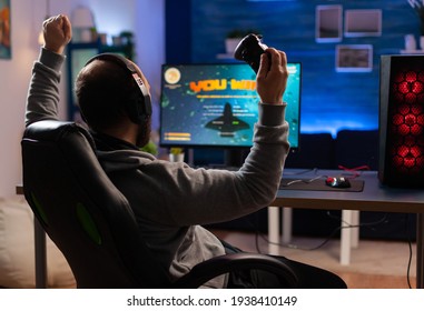 Winner gamer sitting on gaming chair at desk and playing space shooter video games with controller. Man streaming online videogames for esport tournament in room with neon lights