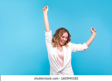 I'm winner! Portrait of ecstatic overjoyed lady with wavy hair in white jacket dancing with raised arms, smiling excitedly, celebrating victory, success. indoor studio shot isolated on blue background