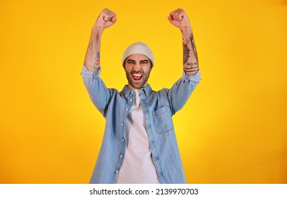 Winner. Rise Up Hands. Studio Shot Of Smiliing Handsome Man In Jeans And White Hat On Yellow Background. Tatoo And Beard. Emotional Concept.