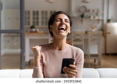 Winner time. Overjoyed young latina female sit on sofa alone hold phone laugh raise up fist proud with victory in game lottery online. Happy excited woman celebrate success scream yes full of emotions - Shutterstock ID 1953915841