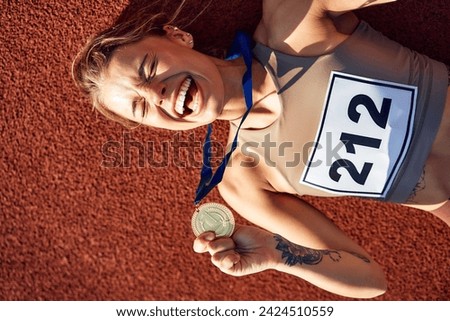 Winner woman in sportswear with number with gold medal for first place and cup lying at stadium celebrating after competition. Sports,  victory concept.
