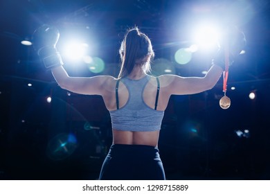 Winner Young female boxer athlete holds gold medal in her hand on dark blurred background, photo from back.