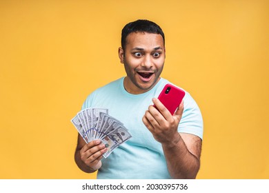 Winner! Young Rich Happy African American Indian Black Man In Casual Holding Money Dollar Bills With Surprise Isolated Over Yellow Wall Background. Using Mobile Phone.