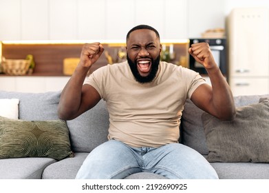 Winning concept. Amazed cheerful young african american man, sitting on the sofa in living room, excited for success with arms raised, celebrating victory, rejoices at a goal, smile happily - Powered by Shutterstock