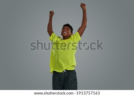 Winning. Excited teenage african boy wearing neon t shirt celebrating success, keeping arms raised while standing against grey background, enjoying victory. Young people and sport