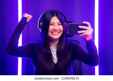 Winning video game on cellphone. Young asian pretty woman sitting on chair holding smartphone in living room. Happy female Pro Streamer chinese wearing headphone playing game online with neon light.