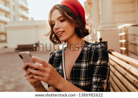 Winsome elegant girl texting message in autumn day. Debonair short-haired woman in red beret standing on the street with phone.