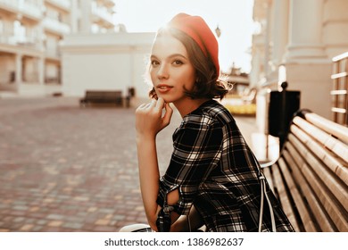 Winsome girl in french beret sitting in park. Outdoor photo of elegant caucasian lady chilling on bench.
