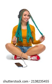 Winsome Positive Caucasian Female with African American Dreadlocks Listens Music in Wirreless Headphones Smartphone While Sitting on Floor Over White. Vertical image