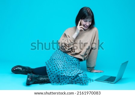Winsome Smiling Caucasian Brunette Girl Posing with Laptop And Cellphone While Surfing The Internet in Hoodie Against Blue Background. Horizontal Orientation