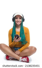 Winsome Smiling Caucasian Female with African American Dreadlocks Listens Music in Headphones On Cellphone Smartphone While Posing in Streetwear Clothing Over White. Vertical image