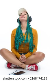 Winsome Smiling Caucasian Female with African American Dreadlocks Listens Music in Headphones On Cellphone Smartphone While Posing in Streetwear Clothing Over White. Vertical Shot