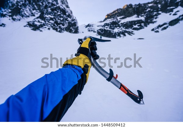 Winter alpine climbing. Hand in\
a winter glove holding an ice axe, snow, ice and rock in the\
background. Point of view of an climbing tool in a hand of an\
alpinist.