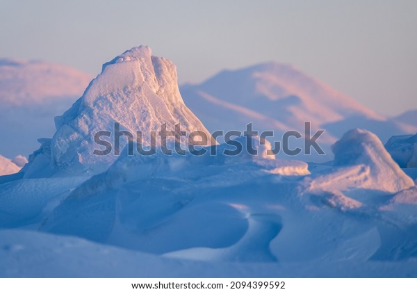 Winter arctic landscape. Ice hummocks and\
snowdrifts against the background of blurred mountains.\
Snow-covered ice floes close-up. Cold frosty winter weather. Polar\
region. Shallow depth of\
field.