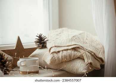 Winter and autumn hygge. Stylish cup of tea with cozy knitted sweater, pine cone, wooden star and golden lights on windowsill in festive scandinavian room. Cozy Christmas. Happy Holidays! - Powered by Shutterstock